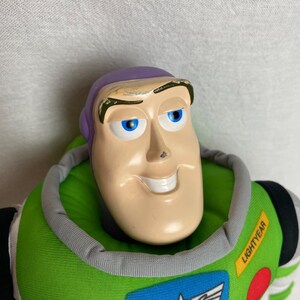 K&ouml;nnte beinhalten: Buzz Lightyear Pl&uuml;schpuppe mit l&auml;chelndem Gesicht, blauen Augen und einem lila Kopf. Die Puppe hat einen gr&uuml;nen und grauen Kragen, und das Wort "Lightyear" ist auf dem gr&uuml;nen Brustbereich aufgedruckt. Die Puppe ist eine Figur aus dem Toy Story-Franchise.