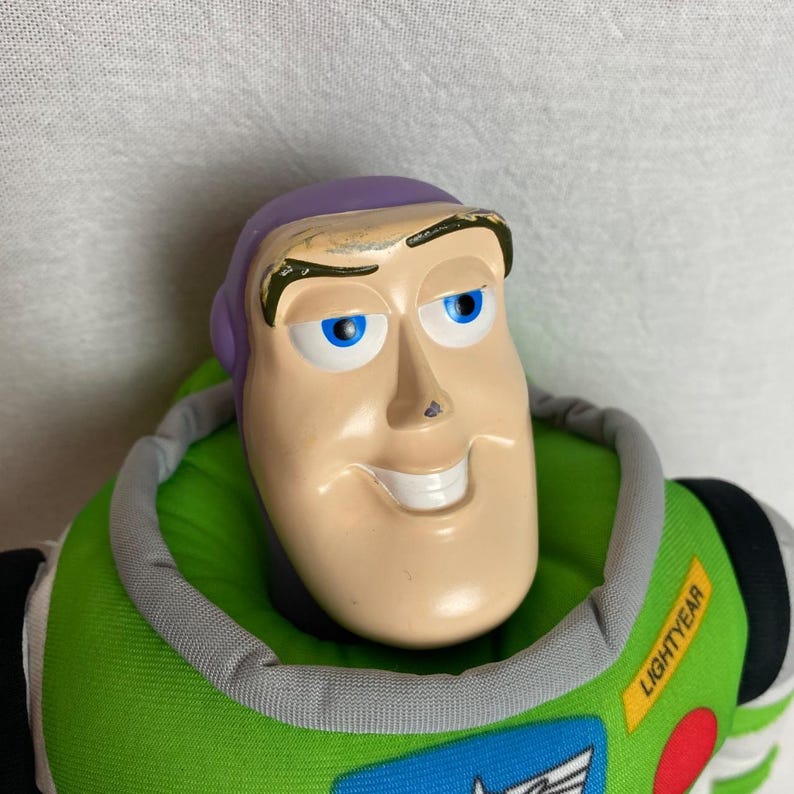 K&ouml;nnte beinhalten: Buzz Lightyear Pl&uuml;schpuppe mit l&auml;chelndem Gesicht, blauen Augen und einem lila Kopf. Die Puppe hat einen gr&uuml;nen und grauen Kragen, und das Wort "Lightyear" ist auf dem gr&uuml;nen Brustbereich aufgedruckt. Die Puppe ist eine Figur aus dem Toy Story-Franchise.