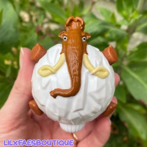 May include: A white, round toy with a brown woolly mammoth design. The mammoth has tan tusks and ears, with a brown trunk and head. The toy has brown feet and is held in a hand. The text "LILXFAESBOUTIQUE" is at the bottom.