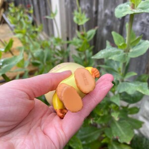 May include: A yellow and orange figurine with the text "Bone Fide" on the bottom of the feet. The figurine is held in a hand, with a blurred background of green foliage and a wooden fence.