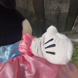 May include: A plush toy character wearing a white glove with three black stitched lines on the back. The character has a black head and is dressed in a shiny pink skirt with a matching pink ruffled sleeve cuff and a light blue satin bodice section.