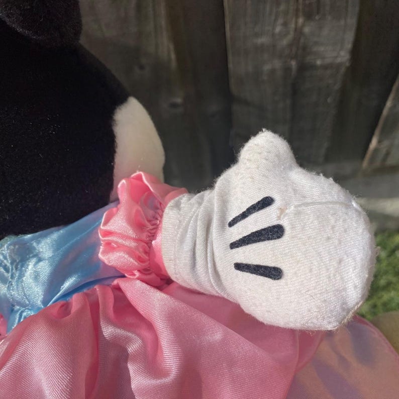 May include: A plush toy character wearing a white glove with three black stitched lines on the back. The character has a black head and is dressed in a shiny pink skirt with a matching pink ruffled sleeve cuff and a light blue satin bodice section.