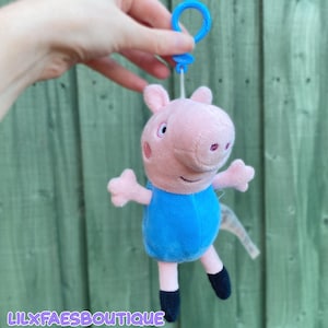 May include: A plush toy of a pink pig character wearing a blue dress, featuring a blue plastic clip attached to the top. The character has small ears, a snout, and black feet. The text LILXFAESBOUTIQUE is printed in white at the bottom of the image.