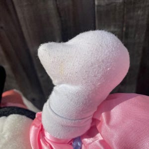May include: A plush toy limb covered in white fabric with a visible cuff at the wrist, attached to a bright pink satin garment. The white fabric shows a slight texture and a small, faint yellowish mark on the upper surface of the rounded hand.