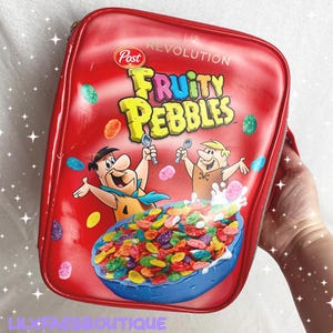 May include: A red rectangular cosmetic bag with a zipper, featuring the Fruity Pebbles cereal logo and cartoon characters. The bag is decorated with colourful cereal pieces. The text "REVOLUTION" and "Post" are also visible.