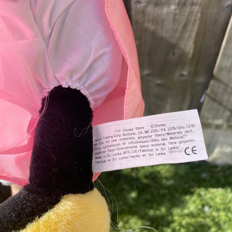 May include: A plush toy featuring a black limb, a yellow foot, and a pink fabric garment with white lining. A white rectangular tag is attached, printed with text including The Disney Store, Disney, and manufacturing details in English.
