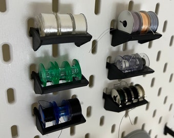 May include: A collection of sewing thread spools in various colors, including white, green, blue, and black, are displayed on black shelves attached to a white pegboard. The spools are filled with thread and are ready for use.