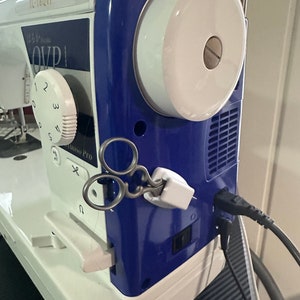 May include: A blue and white sewing machine with a white dial that reads "1, 2, 3, 4, 5, 6" and "OVP" on the side. The machine has a white bobbin winder and a black power cord.