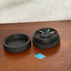 May include: A black plastic storage container with a circular lid and a rotating compartment. The container is open and the compartment is visible. The compartment has a small hole in the center and is divided into four sections. The sections are colored red, white, and blue. A small blue and white memory card is visible in the background.