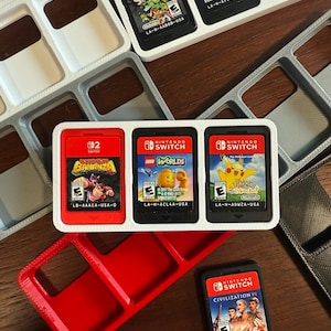 May include: A collection of Nintendo Switch game cartridges and storage cases. The games include titles like "Splatoon," "Lego Worlds," and "Civilization VI." The cartridges are in black cases with colorful game art. The storage cases are in white, gray, and red.