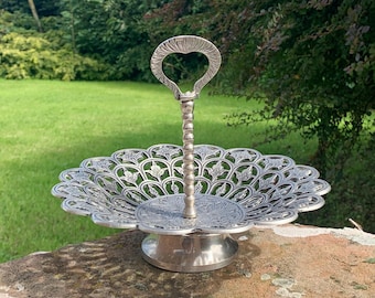 May include: A silver-colored, two-tiered serving dish with a heart-shaped handle. The dish has a decorative, openwork design and sits on a round base. The handle is a vertical rod with a heart-shaped top.