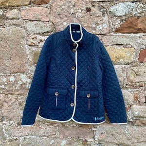 May include: A navy blue quilted jacket with white piping and a button closure. The jacket has two pockets and a small logo on the bottom right side.