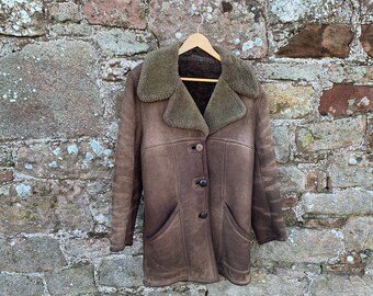 May include: A brown sheepskin coat with a shearling collar and four button fastenings. The coat has two pockets and is hanging on a wooden hanger against a stone wall.