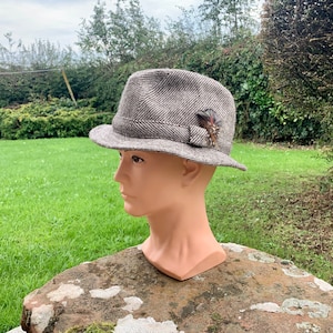 May include: A brown and gray tweed fedora hat with a feather accent. The hat is on a mannequin head.