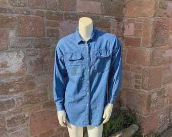 May include: A long-sleeved denim shirt in a medium blue hue. The shirt features two chest pockets with buttoned flaps and a classic button-down front. The collar is a standard point collar. The shirt is displayed on a mannequin.