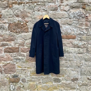 May include: A navy blue long coat with a single breasted closure and a collar. The coat has two pockets on the front and is hanging on a wooden hanger against a stone wall.
