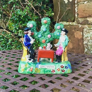 Peut inclure: Figurine en céramique colorée représentant deux personnages assis à une table sous un arbre. Les personnages sont vêtus de costumes d'époque. La base de la figurine porte l'inscription "DR. SYNTAX". La scène se déroule sur une table en métal marron.