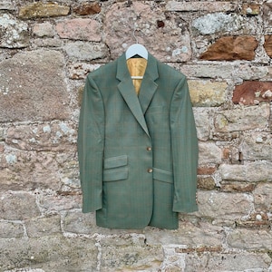 May include: A green plaid blazer with a gold-colored lining hangs on a white hanger against a stone wall. The blazer has two front pockets and two buttons. The fabric has a subtle plaid pattern in shades of green and brown.