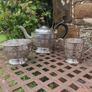 Könnte beinhalten: Ein silbernes Teeservice, bestehend aus einer Teekanne mit schwarzem Griff, einer Zuckerdose und einem Sahnekännchen. Das Set hat ein strukturiertes Design und steht auf einem braunen Metalltisch. Der Hintergrund zeigt Grün und eine Steinmauer.