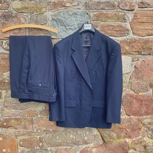 May include: A navy blue pinstripe suit, comprising a blazer and trousers, hangs on a wooden hanger against a stone wall. The blazer has two front pockets and a two-button closure. The trousers are folded neatly.