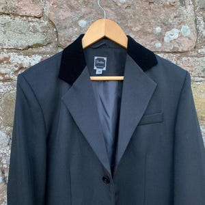 Pode incluir: Um blazer cinza escuro com gola de veludo preto, pendurado em um cabide de madeira. O blazer tem um fecho de um botão e um pequeno bolso no lado esquerdo. O fundo é uma parede de pedra.