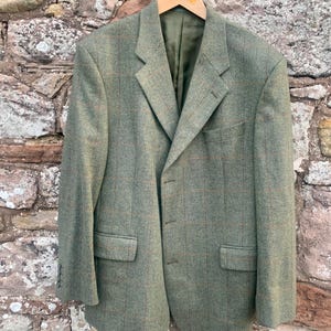 May include: A green tweed blazer with a subtle plaid pattern hangs on a wooden hanger against a stone wall. The blazer has a classic three-button closure, notched lapels, and two front pockets. The lining is a solid olive green.