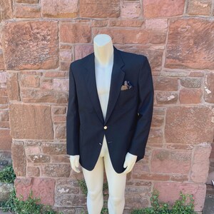 Puede incluir: Un blazer azul marino con un botón dorado y un pañuelo de bolsillo estampado. El blazer se exhibe en un maniquí contra un fondo de pared de ladrillo.