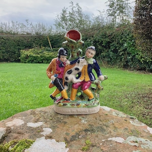 Könnte beinhalten: Keramikfigur, die zwei Figuren in historischer Kleidung zeigt, die ein geflecktes Tier halten. Die Figur hat ein Farbschema aus Rot, Grün, Gelb, Blau und Braun. Auf der Basis der Figur steht "MURDER OF THOMAS SMITH".