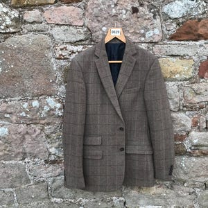 Puede incluir: Blazer de tweed marrón con un sutil estampado de cuadros, colgado de una percha de madera. La chaqueta tiene dos bolsillos delanteros y un forro azul oscuro. El fondo es una pared de piedra.