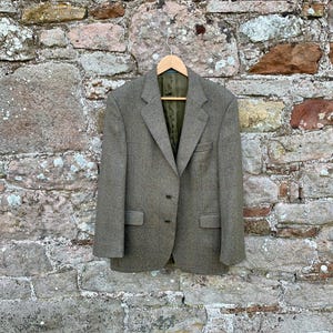 May include: A tweed blazer in shades of olive green and brown hangs on a wooden hanger against a stone wall. The jacket has a classic two-button closure, notched lapels, and two front pockets. The lining is a darker green.