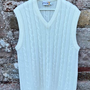 Puede incluir: Chaleco de punto color crema con diseño de trenzas y cuello en V. El chaleco sin mangas cuelga de una percha blanca contra una pared de ladrillos texturizada. La etiqueta dice "PRO TEC".