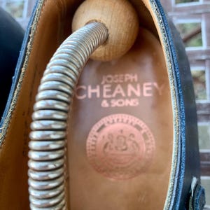 May include: Close-up of a black leather shoe with a wooden shoe tree inside. The shoe's interior is tan, with the brand name "JOSEPH CHEANEY & SONS" printed in copper. A copper emblem is also visible.