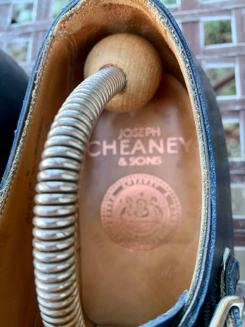 May include: Close-up of a black leather shoe with a wooden shoe tree inside. The shoe's interior is tan, with the brand name "JOSEPH CHEANEY & SONS" printed in copper. A copper emblem is also visible.