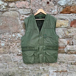 Peut inclure: Gilet vert olive avec plusieurs poches et une fermeture boutonnée. Le gilet a un design matelassé et une doublure foncée. Il est suspendu à un cintre en bois sur un mur de pierre.
