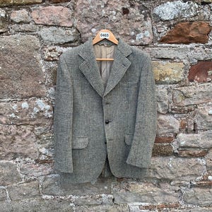 May include: A vintage tweed blazer in a grey and beige houndstooth check, suspended on a wooden hanger. The jacket features a notched lapel, two front pockets, and a three-button fastening. The backdrop is a rustic stone wall.
