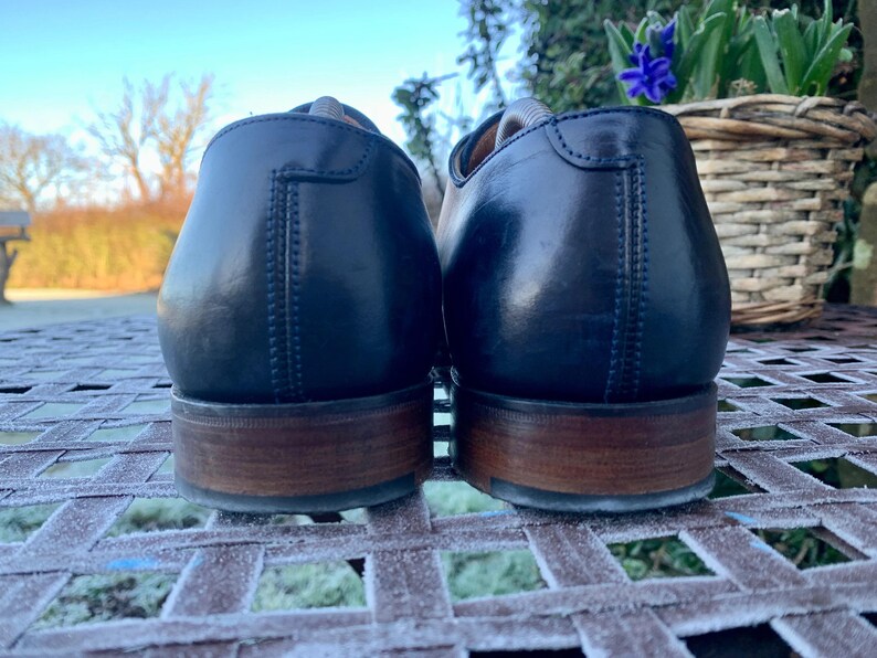 May include: A pair of navy blue leather dress shoes with brown wooden soles. The shoes are positioned close together, showcasing the back of the heels. The image is taken outdoors, with a blurred background of a garden and a clear sky.