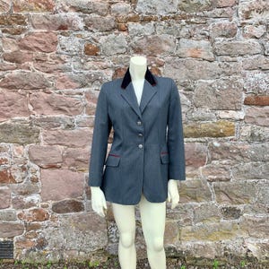 May include: A grey blazer with a black velvet collar and red trim on the pockets and lapel. The blazer has three silver buttons and is displayed on a mannequin. The background is a stone wall.