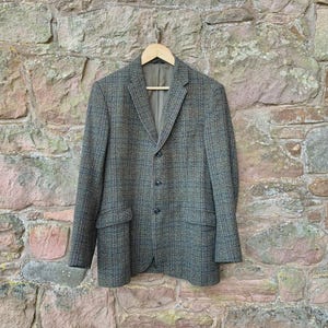 May include: A tweed blazer in shades of gray and blue hangs on a wooden hanger against a stone wall. The jacket has a classic three-button closure, notched lapels, and two front pockets. The fabric has a textured appearance.