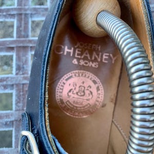 May include: Close-up of a black leather shoe interior with the brand name "JOSEPH CHEANEY & SONS" printed in gold. A wooden shoe tree and metal spring are inside the shoe. The shoe has a buckle closure.