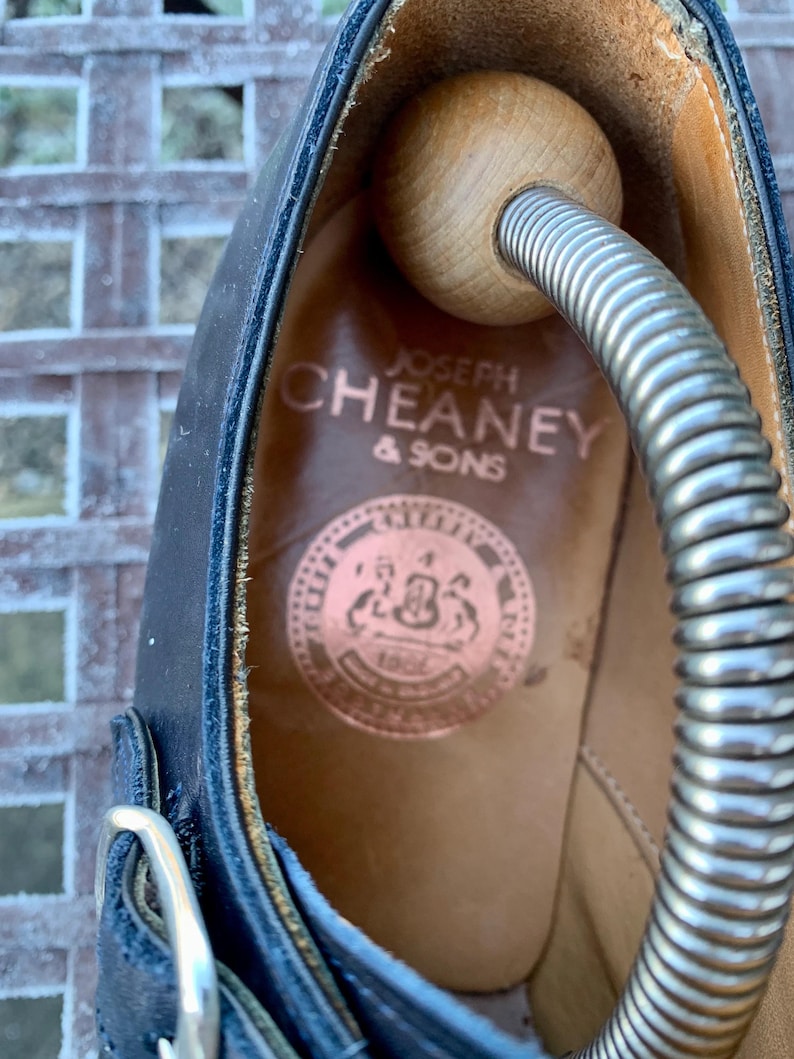May include: Close-up of a black leather shoe interior with the brand name "JOSEPH CHEANEY & SONS" printed in gold. A wooden shoe tree and metal spring are inside the shoe. The shoe has a buckle closure.