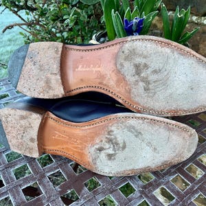 May include: Close-up of the soles of two black leather dress shoes. The soles are worn and show signs of use. The words "Made in England" are visible on one sole. The shoes are on a metal grate.