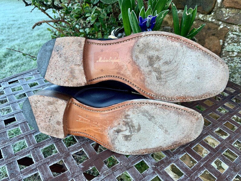 May include: Close-up of the soles of two black leather dress shoes. The soles are worn and show signs of use. The words "Made in England" are visible on one sole. The shoes are on a metal grate.