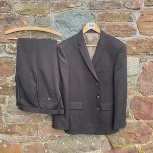 May include: A dark gray suit, including a jacket and trousers, hangs on a wooden hanger against a stone wall. The jacket has three buttons and two front pockets. The trousers are folded neatly. A tag with the number "0649" is attached to the hanger.