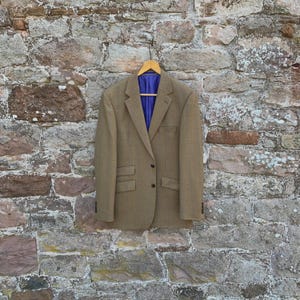 May include: A tan blazer with a blue interior lining hangs on a wooden hanger against a stone wall. The blazer has two buttons and two front pockets. The wall is made of various sized stones.