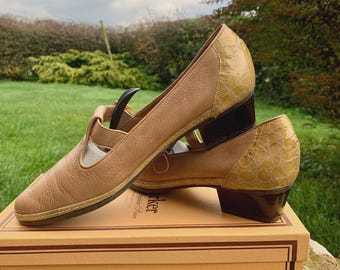 Op de afbeelding: Een paar bruin lederen Mary Jane-schoenen met een getextureerde gele hiel en afwerking. De schoenen staan op een doos met het logo "Barker of Earls Barton". Ze hebben een kleine hak en een bandje.