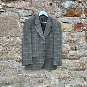 May include: A brown and tan plaid blazer with a single button closure. The blazer is hanging on a white hanger against a stone wall.