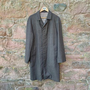 May include: A grey, long coat with a subtle checked pattern hangs on a wooden hanger. The coat has a collared neckline and a button-down front. The coat is displayed against a textured stone wall.