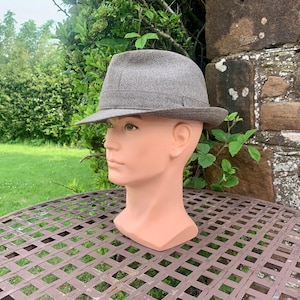 May include: A grey fedora hat with a brown brim sits on a mannequin head. The hat is made of a textured fabric and has a classic design. The mannequin is placed on a metal table outdoors, with a green lawn and stone wall in the background.