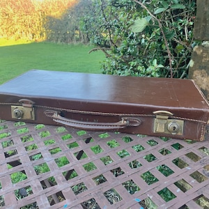 May include: A vintage brown leather briefcase with brass locks and a handle. The rectangular case is closed and sits on a metal table outdoors. The briefcase appears to be well-worn.