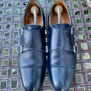 May include: A pair of navy blue leather double monk strap shoes with silver buckles. The shoes have wooden shoe trees inside. The word "CHEY" is visible on the inner lining of one shoe.